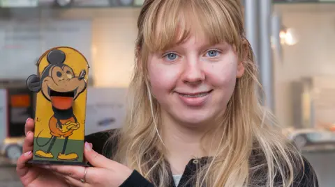 David Duggleby Auctioneers A young woman with blonde hair and blue eyes holds an old money box bearing the image of Mickey Mouse.