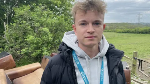 BBC/Seb Cheer A young man with blond hair stands outside, looking into the camera lens. He is wearing a grey hoodie and navy coat, as well as a light blue lanyard which says "We're Kirklees" on it. Behind him is a pile of brown sofas, and a luscious green landscape in the background.