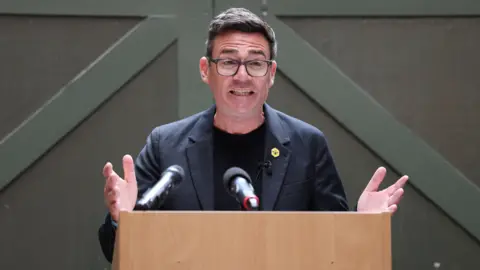 Andy Burnham mid-speech, wearing glasses, suit and tie, with arms stretched out and open palms.