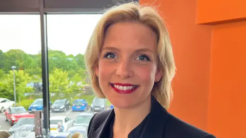 Woman with short blonde hair red lipstick black blazer stood in front of a window 