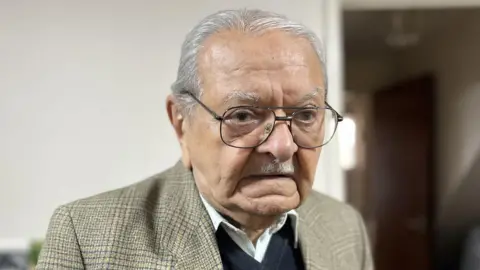 A portrait image of Mr Osman. He is 91 and wearing a brown/grey checked blazer with a dark blue swearer and revealing the collars of a white shirt. 