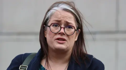 PA Media A woman, pictured from the shoulders up, with brown and grey hair. She is wearing a navy coat and glasses. 