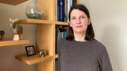 Helen stands in front of a bookshelf, wearing a brown sweater. She has brown, shoulder-length hair and brown eyes. Various figurines and pictures adorn her wooden bookshelf as well as several art books.