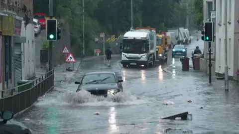 A car driving through flood water, half submerged. There is some rubbish floating in th water.