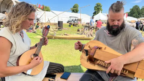 Mason and Mike sitting down on a bench facing towards each other. Mason is playing a wooden mandolin while Mike is playing a hurdy-gurdy, which is also wooden and has a number of small keys which he is pushing with one hand and a crank he is turning with the other.