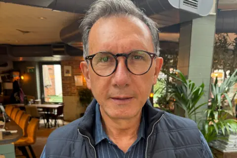 Murat Askin, wearing glasses and a jacket, looking at the camera inside a restaurant.