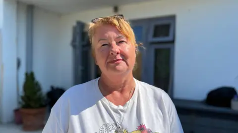 Annette is a woman with light strawberry blonde hair. She is wearing a white t-shirt and has glasses on her head. She has a silver necklace around her neck. She is standing in her garden, in front of the backdoor to her living room. There is sunshine on her face and she is looking into the camera.