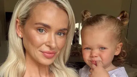 Robyn Baker has long blonde hair. She is holding her daughter on her left, who has short brown hair tied in a top-knot.