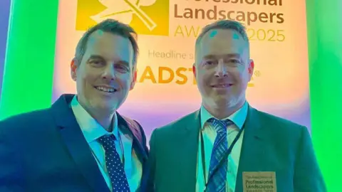 Native Landscape Design Mike Walker and Matthew Stevenson, both wearing smart suits and ties, pose together at an award ceremony in front of a sign saying Professional Landscapers Awards