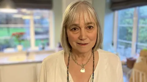 Penny Parry is standing in her kitchen wearing a white shirt. She is smiling.