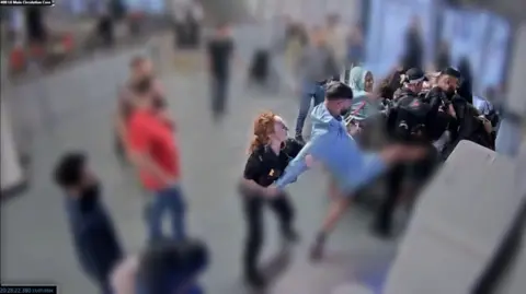PA Media A man in blue kicks in the direction of a police officers while a police officer with red hair holds his arms behind him at the car park pay stations at Manchester Airport.
