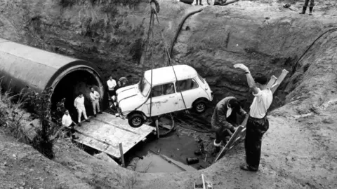 Coventry Telegraph Archive/Mirrorpix/Getty Images A Mini is lowered into Coventry sewers during the filming of The Italian Job film. 26th September 1968. A man stands next to a large hole with his arm out, hand down, to indicate the direction of the winching. Several people stand next to a pipe looking up at the Mini in the air