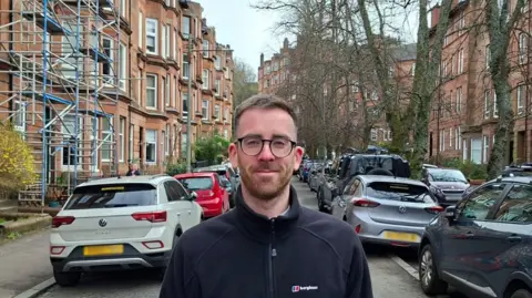 david  kelly wearing glasses and a dark outdoor jacket stands in the foreground of a residential street lined with parked cars and red-brick buildings. Scaffolding is visible on one building to the left, and trees without leaves line the street in the background.