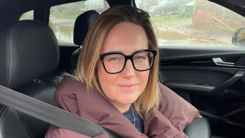 John Devine/BBC Emma Watts has sandy/brownish hair just down to her shoulders, she is wearing a light purple coat over a blue shirt with a collar, she is sitting in the driving seat of her car with a seat belt on.