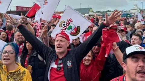 BBC Saints fans wave flags and hold their arms in the air outside St Mary's Stadium