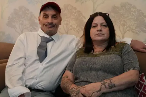 A man and a woman sit next to each other on a brown sofa with grey wall paper showing a tree pattern behind. Both are middle aged. The man is wearing a red baseball cap and has a moustache and a nose-ring. He is wearing white shirt with a blue-grey tie, tucked inside his shirt. The woman has shoulder length reddish-brown hair and has glasses on top of her head. She is wearing a grey and green short sleeved top and has tatoos on her forearms. Both are looking directly at the camera.