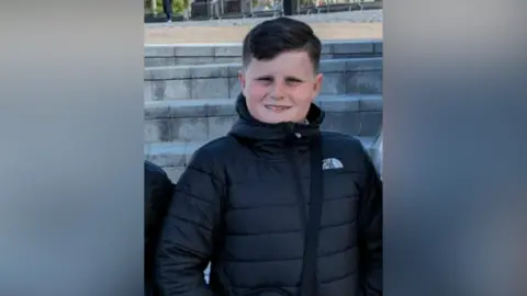 Trainor family A young boy smiles at the camera, he is wearing a black anorak and has short, black hair which is swept to one side. He is outside, the background appears to be a town square. 