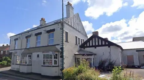 A Google Street View screenshot of the Royal Hotel.  The large two-storey building sits at the intersection of two streets. There is an empty concrete yard to the side with overgrown vegetation and some rubbish.