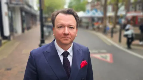 Michael Keohan/BBC A man with dark blond hair, wearing a navy suit and a black tie with small red dots, is standing at a High Street.