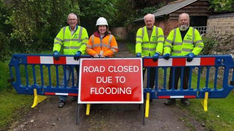 Four volunteers in hi-vis jackets with a flood barrier and sign