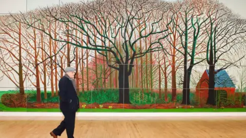 A man wearing a hat and scarf is walking in front of a very large painting depicting trees 