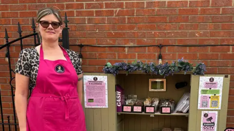 Heather Price in a pink apron and sunglasses standing next to her cake shed. The doors are open and several cakes are on display.