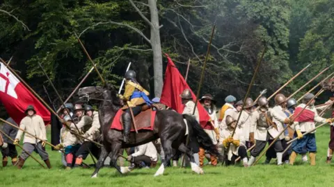 Sealed Knot A civil war enactment taking place, showing two horses running, a large number of people dressed as soldiers, in uniform, holding sticks. They are outside, with trees behind them.