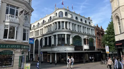 A department store named Jarrolds stands alongside empty roads as pedestrians walk past on a summer evening with clear blue skies