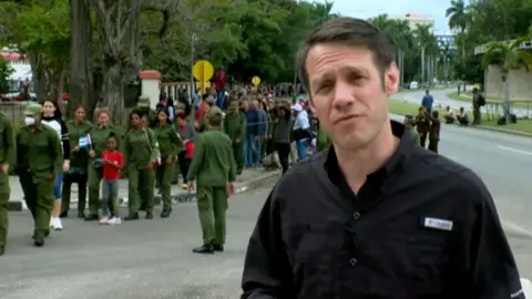 BBC correspondent Will Grant on Cuban street with army personal in the backgroud.