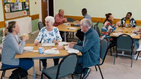An older man and woman sat at a table next to a younger woman in a community centre. They all have colourful stripey mugs and are smiling.