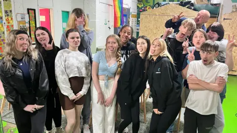 A group of 12 young people stand with a youth worker. They are making funny expressions for the camera. 