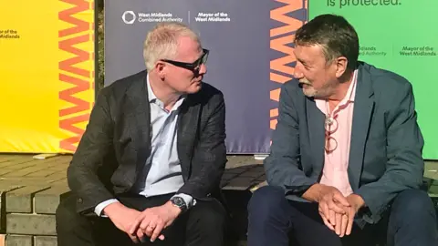 LDRS West Midlands Mayor Richard Parker and Steven Knight sat on a brick wall in front of hoardings, both wearing suits and speaking to each other