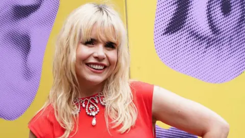 Paloma Faith smiles, she has bleach blonde wavy hair. She wears an orange tope and elaborate pearl and diamond necklace. She stands in front of a yellow and purple background.