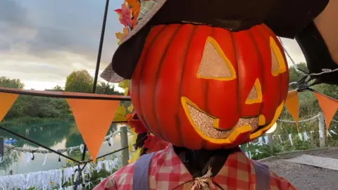 A scarecrow with a  pumpkin for a head is standing in front of a fishing lake