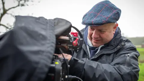 Daniela Cotognini Mick, wearing a flat cap and a rain coat, looks at a camera. He is standing outside in the countryside.