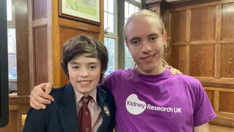 A teenage boy with dark brown hair wearing a maroon tie and red and white striped shirt and school blazer being hugged by an older boy with long, blond hair and wearing a purple t-shirt 