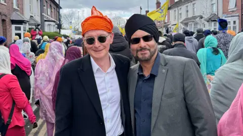 Jules Singh A white man in a black suit and a white shirt and orange scarf around his head smiles next to a man in a grey suit, blue shirt and black turban. Both are wearing sunglasses and behind them are hundreds of people watching a procession 