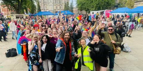 Preston City Council A large crowd of people on Preston's Flag Market, looking and pointing at the camera and smiling, wearing casual clothing and some with rainbow pride flags