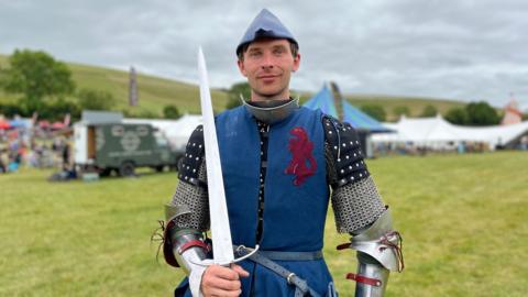 A man looking at the camera in armour with a blue jacket holding a sword