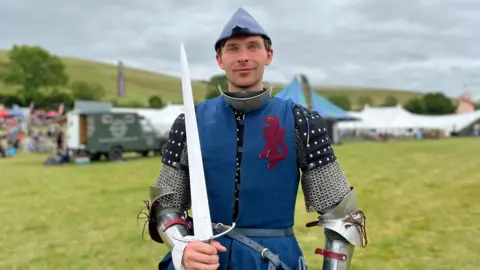 BBC A man looking at the camera in armour with a blue jacket holding a sword