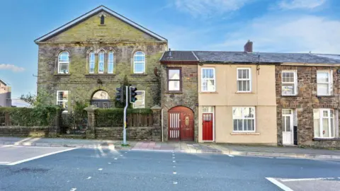 Paul Fosh Auctions A general view of property on Llewellyn Street. It is a brick house with a wooden door and window frame. The house sits on the main street in front of a set of traffic lights. 