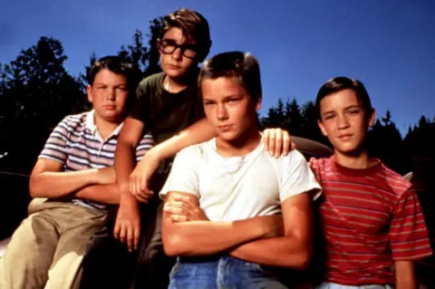 Everett/Shutterstock Jerry O'Connell, Corey Feldman, River Phoenix and Wil Wheaton standing next to each other with arms folded and draped on each other in front of trees and a blue sky