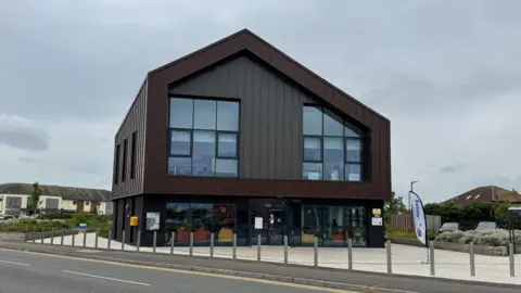 A photograph of the outside of the Campus for Future Living building in Mablethorpe. It is a modern-looking building with large glass windows, brown cladding and an asymmetrical roof. The building is surrounded by smart, plaza-style paving.