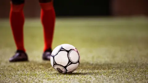 Getty Images A green football pitch. In the centre there is a football. Behind the football are two legs, from the knees down, wearing red knee high socks and black football shoes. 