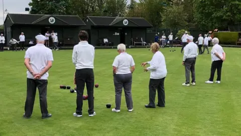 Longthorpe Bowls Club Members f the club playing bowls - wearing white shirts and black trousers, standing on the green grass.