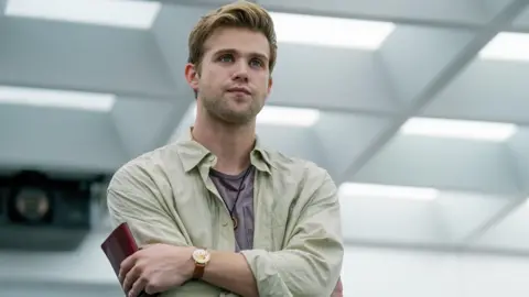 Apple TV+ Leo Woodall acts in Prime Target. He holds a red passport in his hand as his arms are crossed, looking like he is in an office building with skylights. He wears a brown old-fashioned watch and a casual open shirt over a lilac t-shirt.