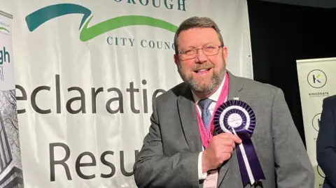 BBC Chris Harper with a beard and glasses wearing a grey suit. He is touching his purple and white rosette with his name on it. A Peterborough City Council sign saying "Declaration Results" is in the background.