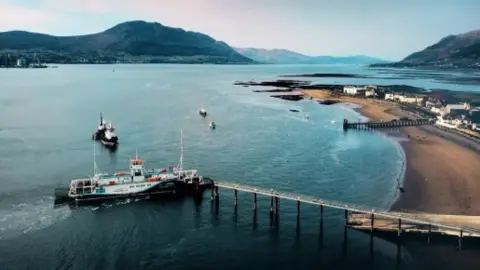 An aerial view of the Carlingford Lough Ferry coming into dock at Greencastle, County Down. A long pier stretches into the water in front of a long, sandy shore. Houses and small buildings line the beach. There are mountains hugging the coastline around either side of the lough.