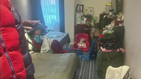 A room with a striped carpet and two beds. A boy in a wheelchair can be seen. His face is blurred/