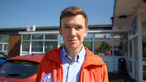 Dan Jones looks at the camera. He has short brown hair and is clean shaven. He is wearing a blue shirt and orange mountain rescue jacket. Behind him is a single storey hospital building
